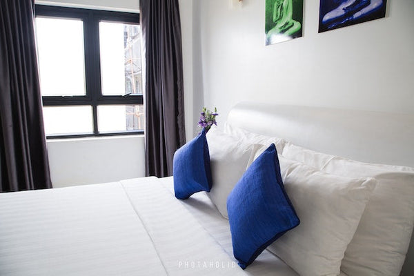 A bed with crisp white linens and blue cushions, by a sunny window