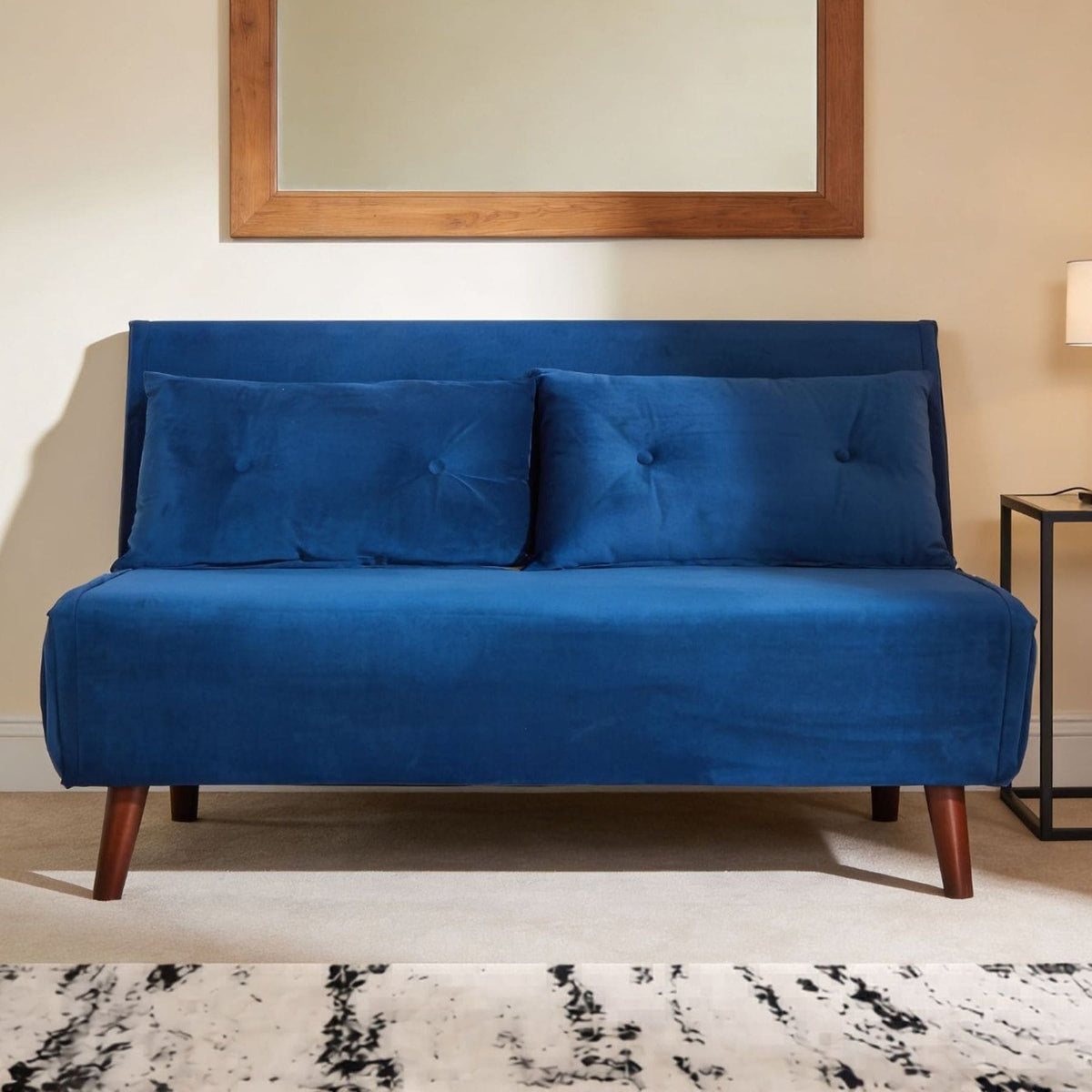 A blue velvet futon sofa with a high tufted backrest and two matching pillows, shown in a styled room setting with a wooden-framed mirror, a side table, and a patterned rug.